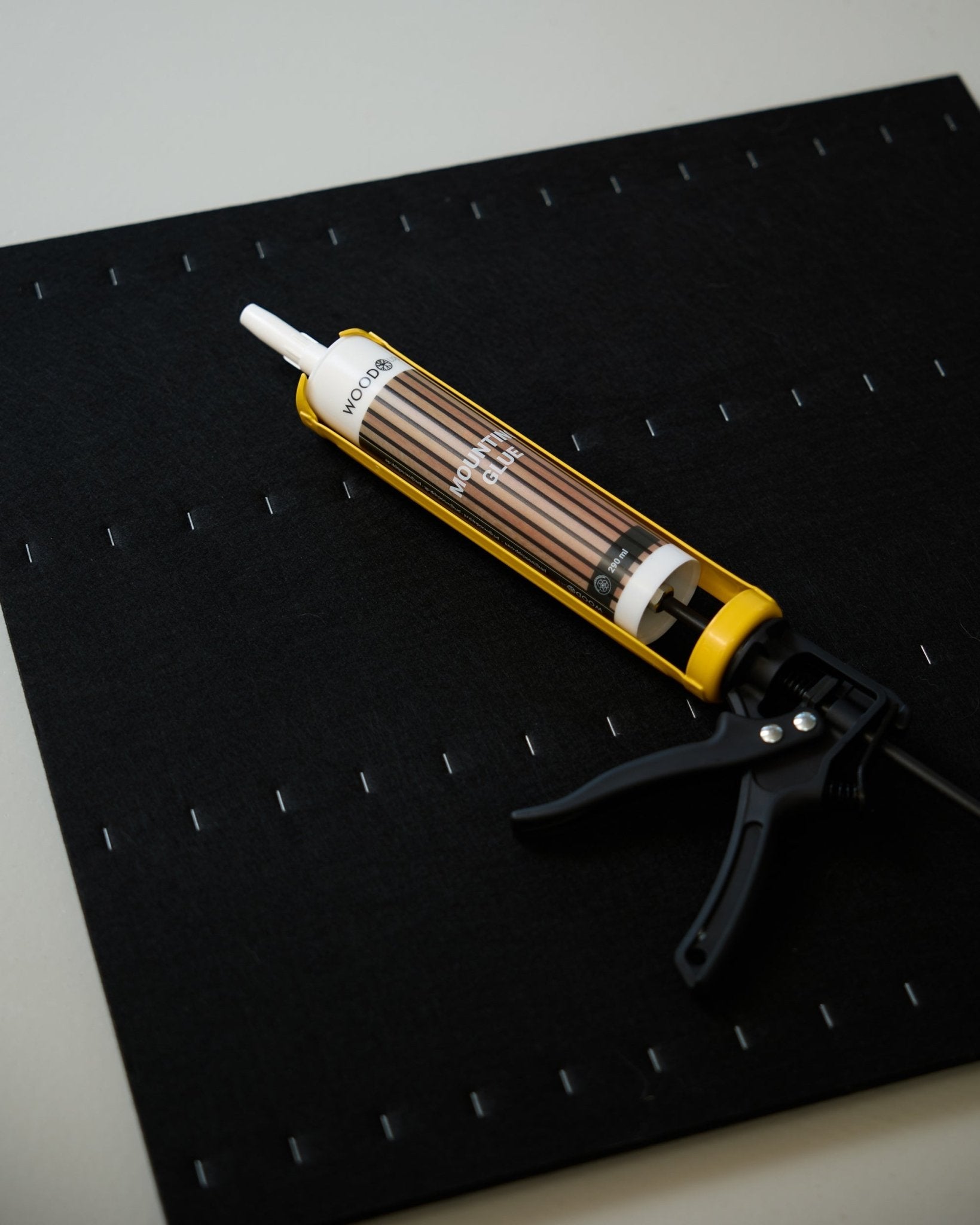 En gul og black WoodUpp fugemassepistol (290 ml) ligger på en black matte med et rutenett av hvite stifter, brukt til montering av akustiske paneler.